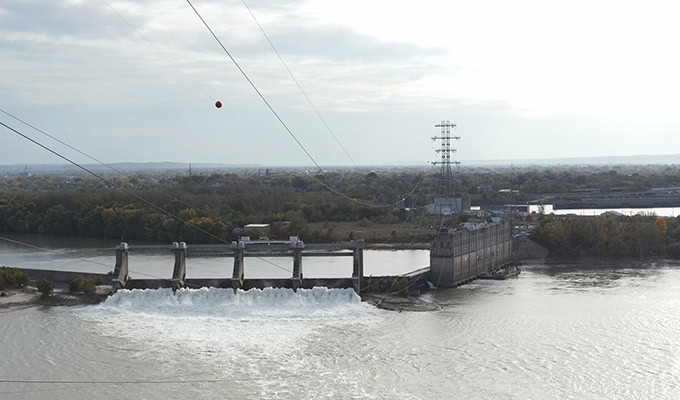 A large concrete dam at Ohio Falls releases water through its spillways, with River Crossing Towers and power lines overhead, set against a backdrop of trees and distant infrastructure.