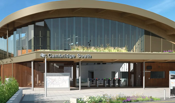 Modern train station entrance with a curved wooden roof, large glass windows, and a sign reading "Cambridge South." Ticket barriers and a map are visible at the entrance.