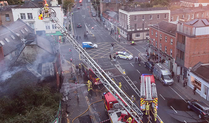 Firefighters use a ladder truck to respond to a building fire near a busy intersection, with smoke visible and emergency vehicles on the street.
