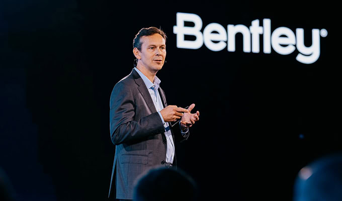 A man in a suit gives a presentation on stage with the Bentley logo displayed on a screen behind him.