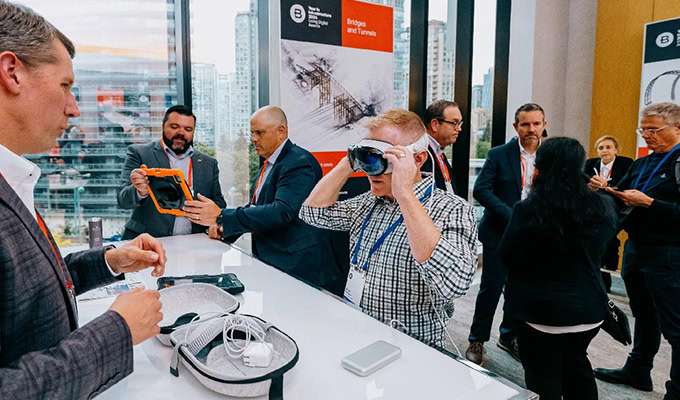 A group of people at a tech event; one person tries on a VR headset at a demonstration table while others converse and examine devices.