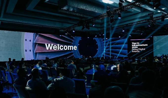 A large conference room with attendees seated facing a stage displaying “Welcome” and “Year in Infrastructure 2024 Going Digital Awards” on large screens.