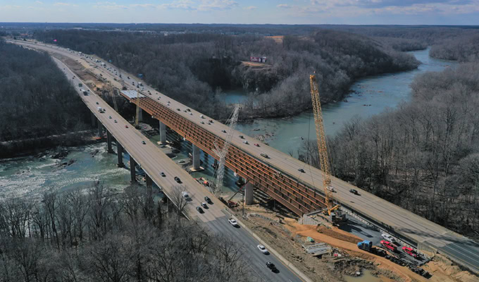 Aerial view of the I-95 Rappahannock River Crossing construction project beside an existing highway bridge over a river, with vehicles traveling on both bridges and construction equipment visible.