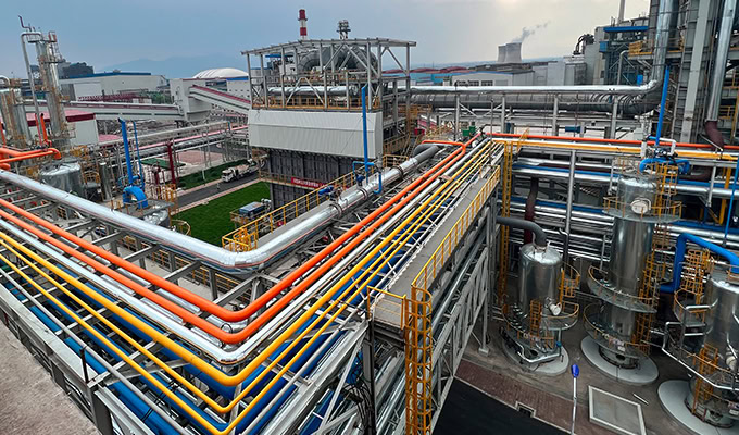 Industrial facility with numerous large metal pipes, tanks, and structures; some pipes are color-coded orange, blue, and yellow. The setting appears clean and organized.