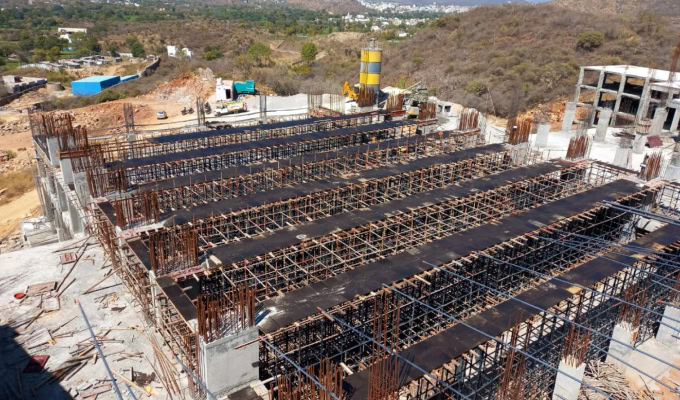 A construction site showing the framework and scaffolding for a multi-story concrete building in progress, with hills and distant structures in the background, reminiscent of the scenic views near Fairmount Palace Udaipur.