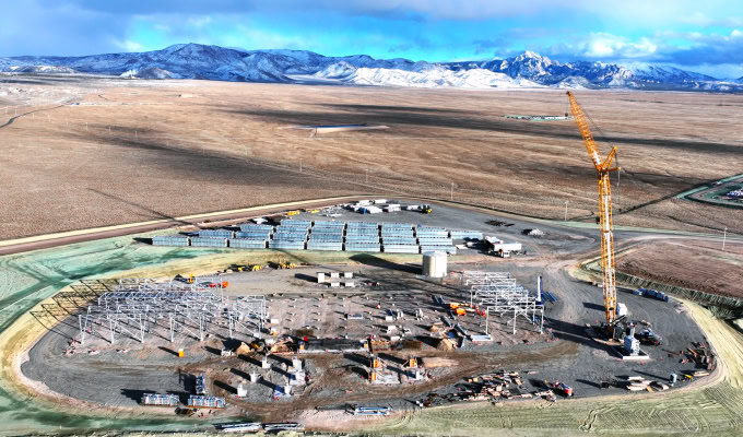 A construction site with steel framework, building materials, and a large yellow crane rises at Cape Station, set against an open, arid landscape with mountains in the background.