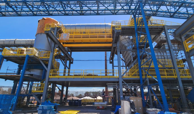 Industrial facility with large metal structures, yellow railings, and blue supports under a clear sky, possibly part of an iron ore briquetting or processing plant using SYNCHRO 4D technology.