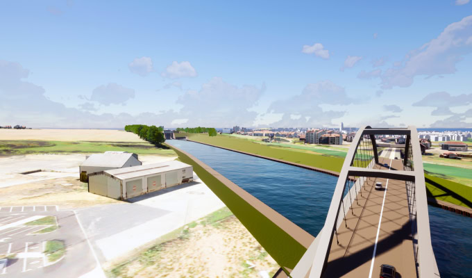 A bridge crosses over the wide Canal Seine Nord Europe with cars driving on it; buildings, a parking lot, and open land are visible on both sides under a blue sky.