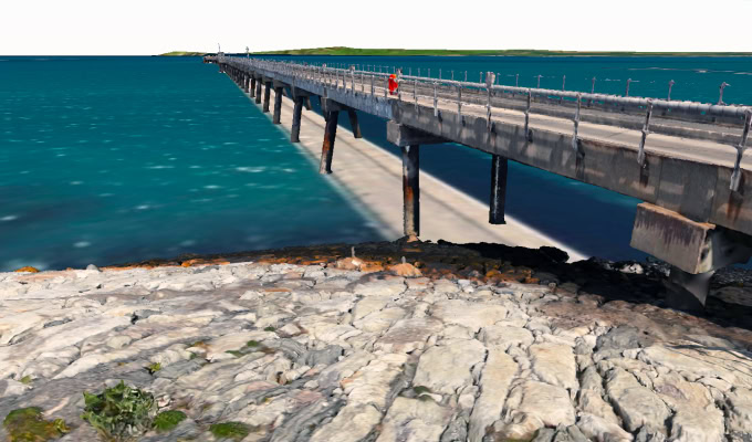 A person in red stands on the Deepwater Jetty at Holyhead, extending over turquoise water, with rocky shore in the foreground and distant land visible on the horizon, suggesting an inspection of the scenic coastline.