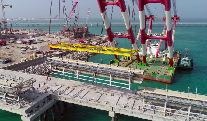 A large crane on a barge lifts a precast concrete structure onto a bridge under construction over turquoise water, with construction equipment and workers nearby.