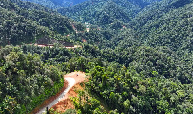 Aerial view of the Sarawak Sabah Link Road, Phase 2, cutting through a densely forested mountainous area, with lush green trees and hills surrounding the cleared path.