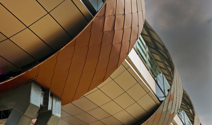 Curved modern building facade with bronze and white paneling, glass windows, and exposed structural elements forms a striking addition to the New Delhi Railway Station Skywalk against a cloudy sky.