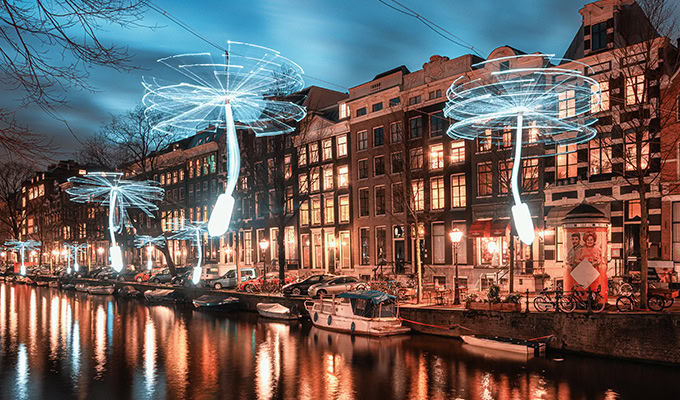 An illuminated art installation resembling giant dandelions is suspended above a canal lined with historic buildings and parked boats at dusk.