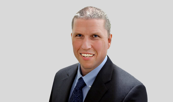 Ein Mann mit kurzen grauen Haaren, einem dunklen Anzug, einem blauen Hemd und einer blauen Krawatte, lächelt vor einem schlichten hellgrauen Hintergrund in die Kamera.