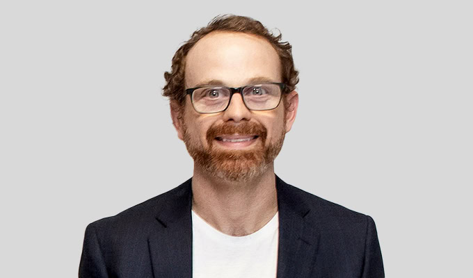 Ein Mann mit Brille, kurzem Bart und lockigem Haar lächelt, während er vor einem schlichten hellen Hintergrund einen dunklen Blazer über einem weißen Hemd trägt.