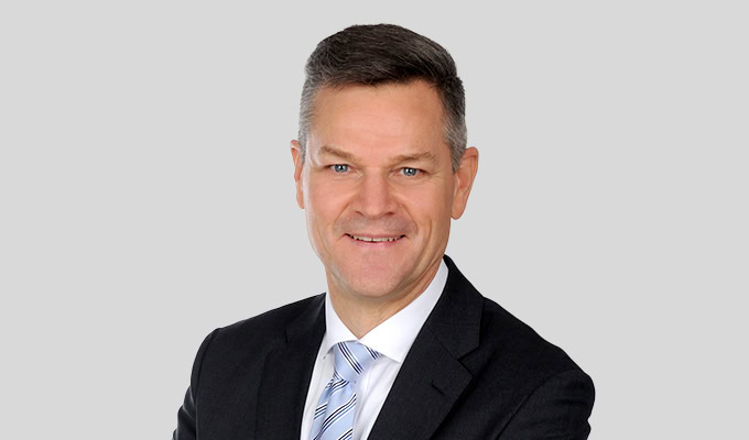 Ein Mann in einem dunklen Anzug, einer hellblau gestreiften Krawatte und einem weißen Hemd posiert lächelnd vor einem schlichten grauen Hintergrund.