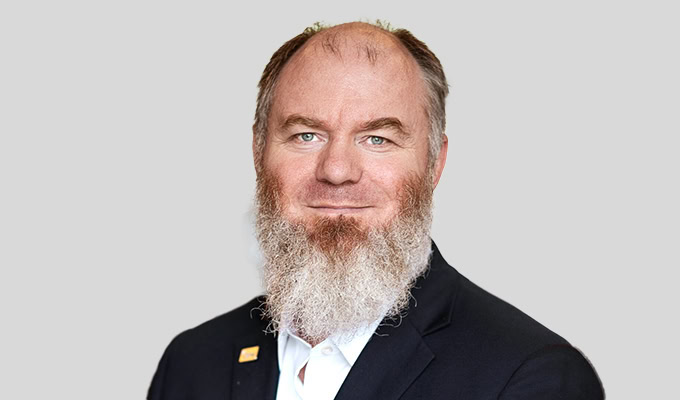 Ein Mann mittleren Alters mit Vollbart, einem schwarzen Jackett, einem weißen Hemd und einer Anstecknadel posiert vor einem schlichten hellgrauen Hintergrund.