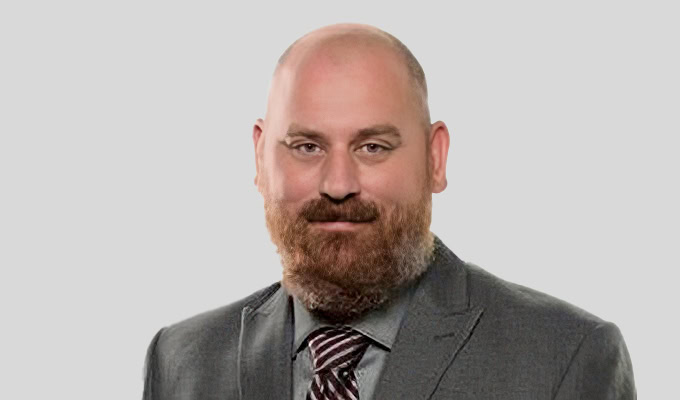 Ein kahlköpfiger Mann mit Vollbart, der einen grauen Anzug, ein Hemd und eine gestreifte Krawatte trägt und vor einem schlichten hellgrauen Hintergrund posiert.