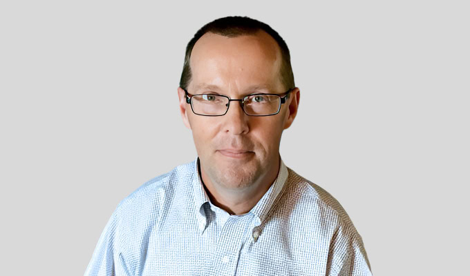 Ein Mann mit kurzen Haaren und Brille, der ein hellblaues kariertes Hemd trägt, posiert vor einem schlichten grauen Hintergrund.