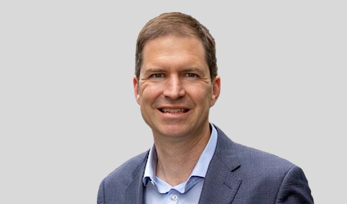 Ein Mann mit kurzen braunen Haaren, einem blauen Jackett und einem hellblauen Hemd, lächelt vor einem schlichten hellgrauen Hintergrund.