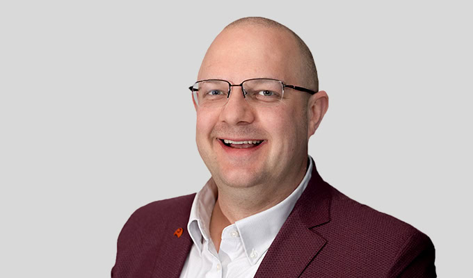 Ein lächelnder Mann mit Brille trägt einen kastanienbraunen Blazer und ein weißes Hemd und posiert vor einem schlichten hellgrauen Hintergrund.