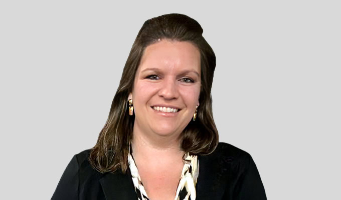 Eine Frau mit langen braunen Haaren, einem schwarzen Blazer und einer gemusterten Bluse, posiert lächelnd vor einem schlichten hellgrauen Hintergrund.