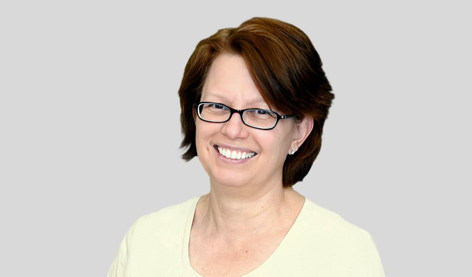 Eine Person mit kurzen braunen Haaren und Brille lächelt und trägt ein helles Oberteil vor einem schlichten hellgrauen Hintergrund.