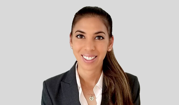 Eine Frau in einem dunklen Blazer und einer weißen Bluse lächelt vor einem schlichten hellgrauen Hintergrund in die Kamera.