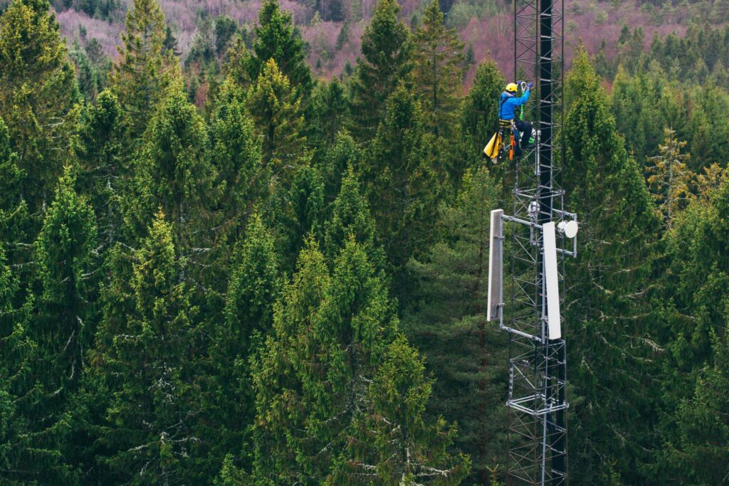 A worker in safety gear climbs a tall communication tower surrounded by dense evergreen forest.