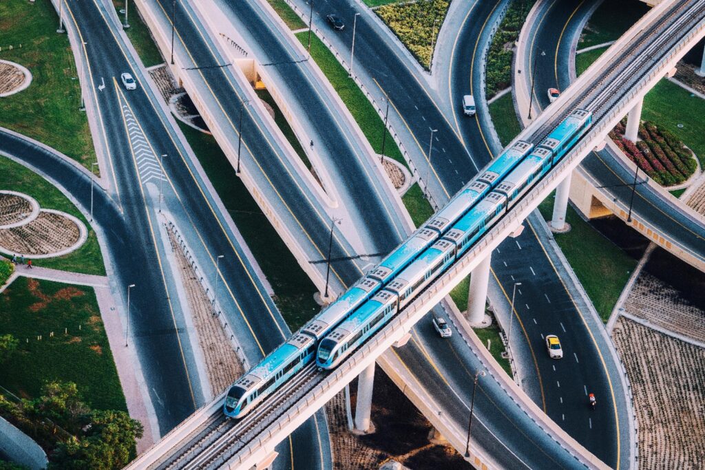 A blue train travels on an elevated track above a network of multilane highways and overpasses, with cars moving below.