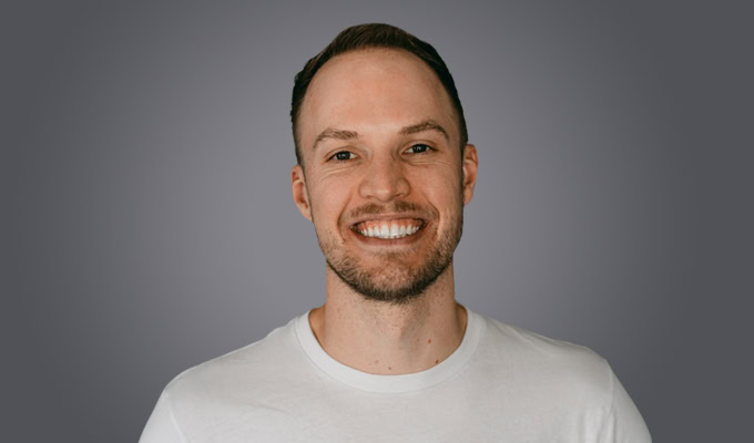 Mark Pittman, a man with short brown hair and a trimmed beard, smiles at the camera in a white t-shirt against a plain grey background.