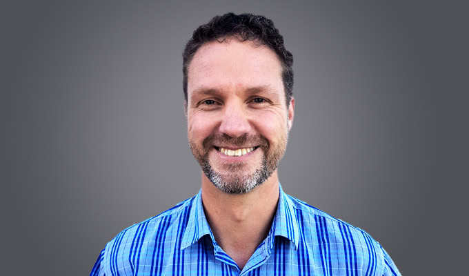 Man with short curly hair and a beard smiling, wearing a blue plaid shirt, in front of a plain gray background.