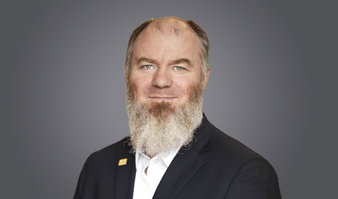 A middle-aged man with a bushy gray beard, Andy Kaiyala wears a dark suit jacket and white shirt, posing against a plain gray background.