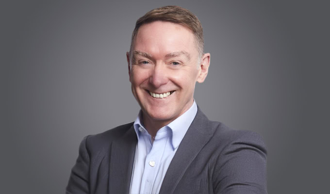 David Lawson, dressed in a suit jacket and light blue shirt, smiles at the camera against a plain, gray background.