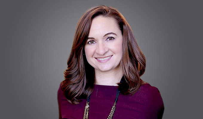 Kristin White, a woman with long brown hair, wearing a maroon top and a necklace, smiles at the camera against a gray background.