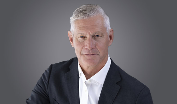 A middle-aged man with short gray hair wearing a dark suit jacket and white shirt poses in front of a plain gray background.