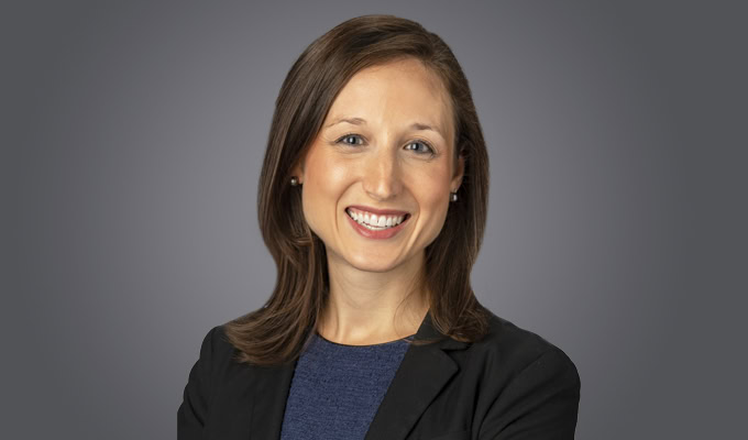 Stephanie Gossman, with straight brown hair, wears a dark blazer and blue top, smiling at the camera against a plain grey background.
