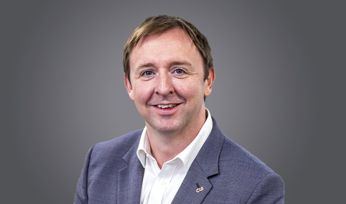 Graham Openshaw, a man with light brown hair in a blue blazer and white shirt, smiles at the camera against a plain gray background.