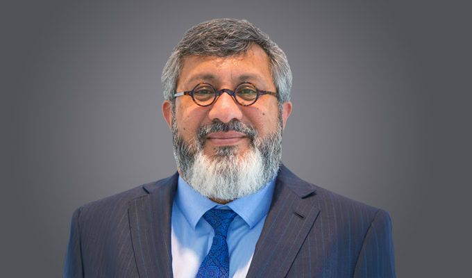 A man with glasses and a gray beard, wearing a dark suit, light blue shirt, and patterned blue tie, poses against a plain gray background.