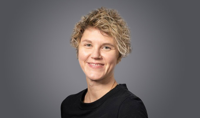 Vicky Ridley-Pearson, with short, curly blond hair and a black shirt, smiles in front of a plain gray background.