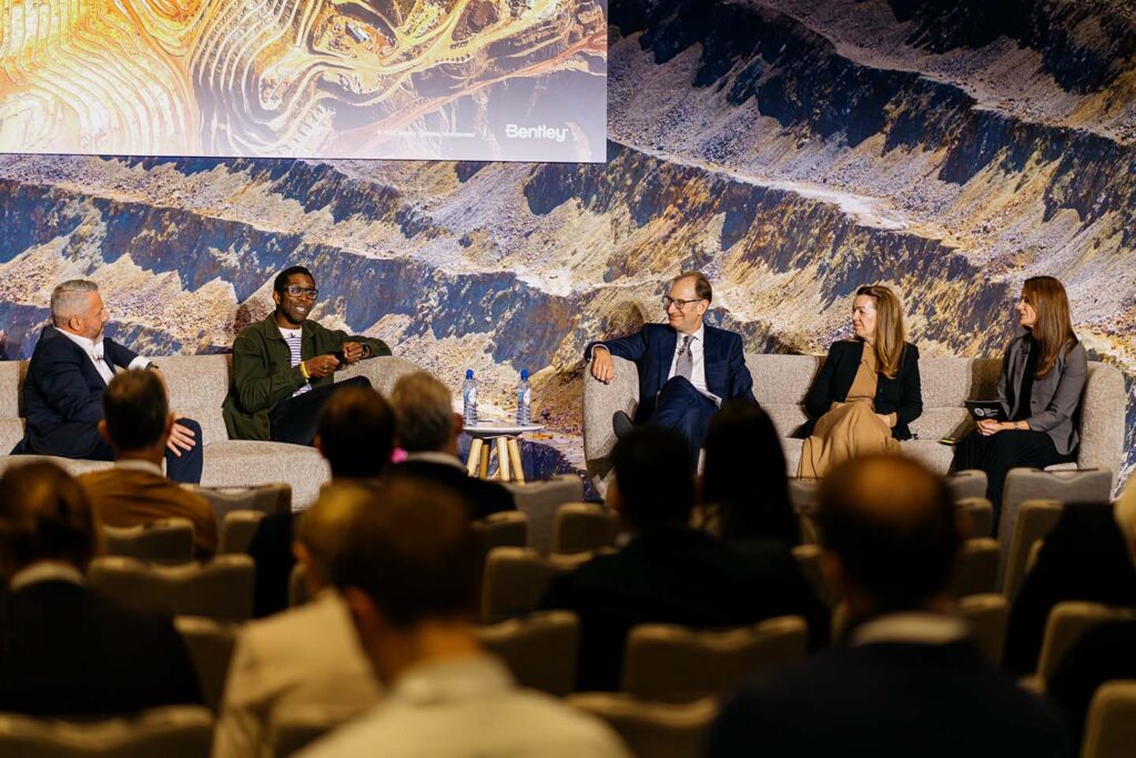 Five panelists sit on stage having a discussion in front of an audience, with a large mining-themed backdrop behind them.