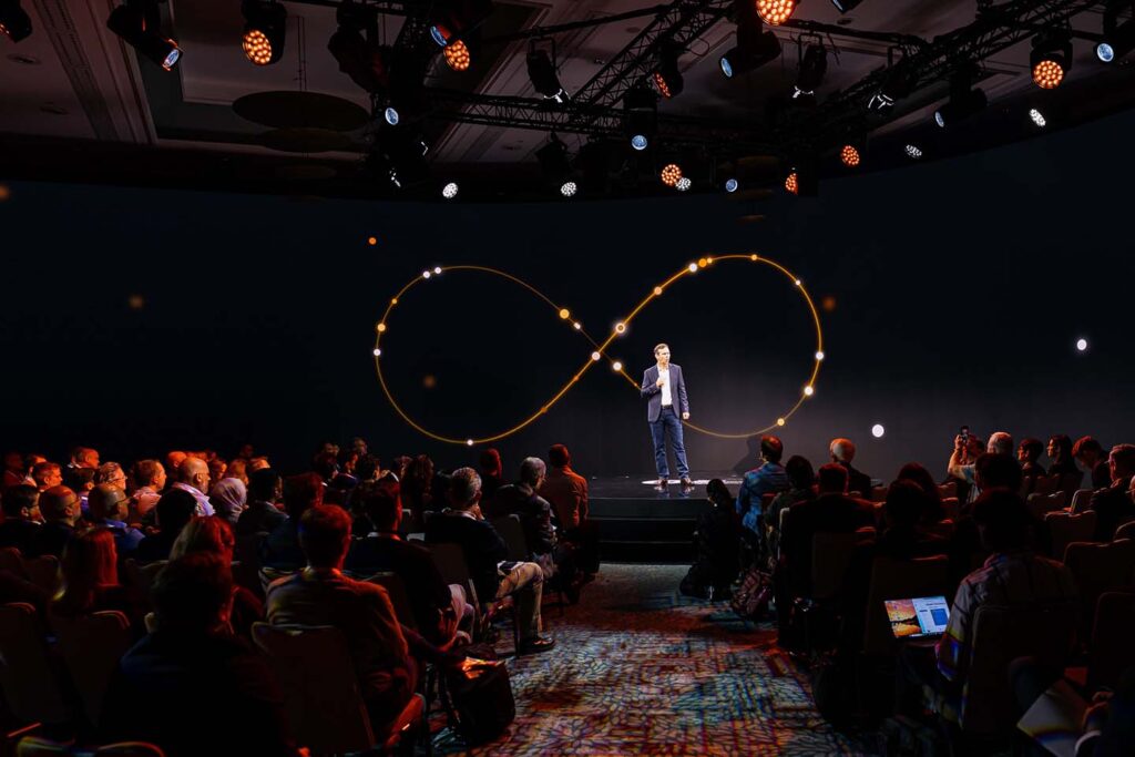 A speaker stands on stage in front of an infinity symbol projected on a screen, addressing an audience seated in a dimly lit conference room.