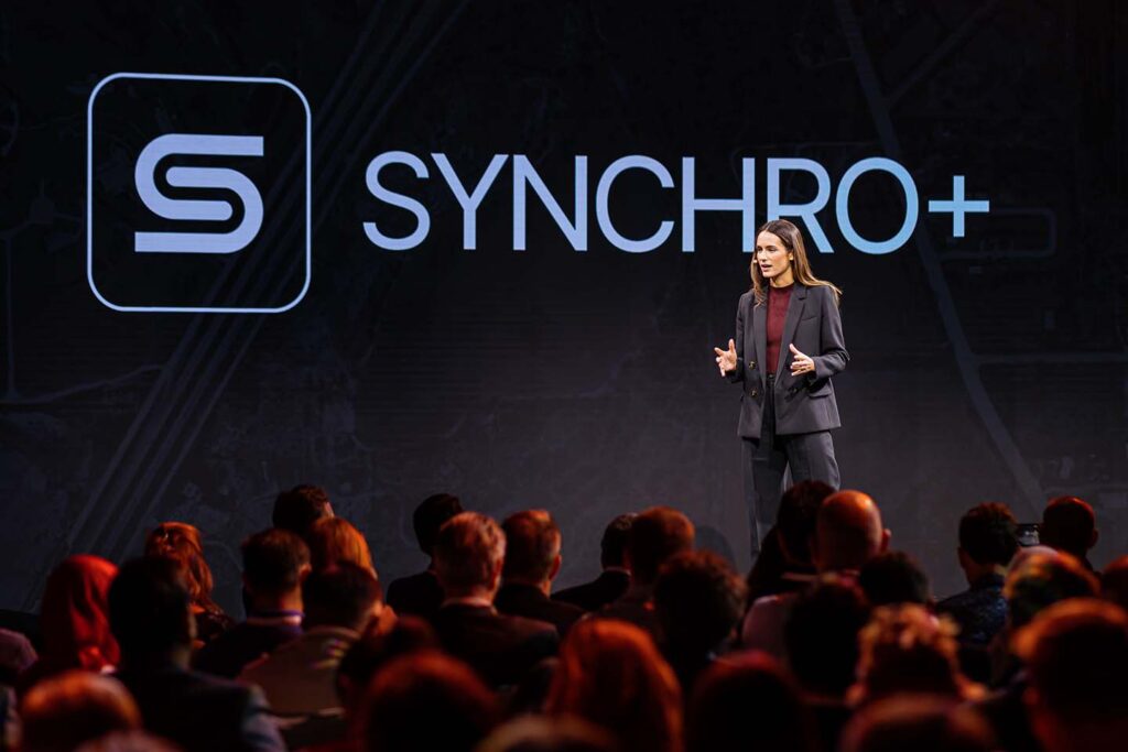 A woman in a suit presents on stage in front of a seated audience, with the Synchro+ logo and name displayed on a large screen behind her.