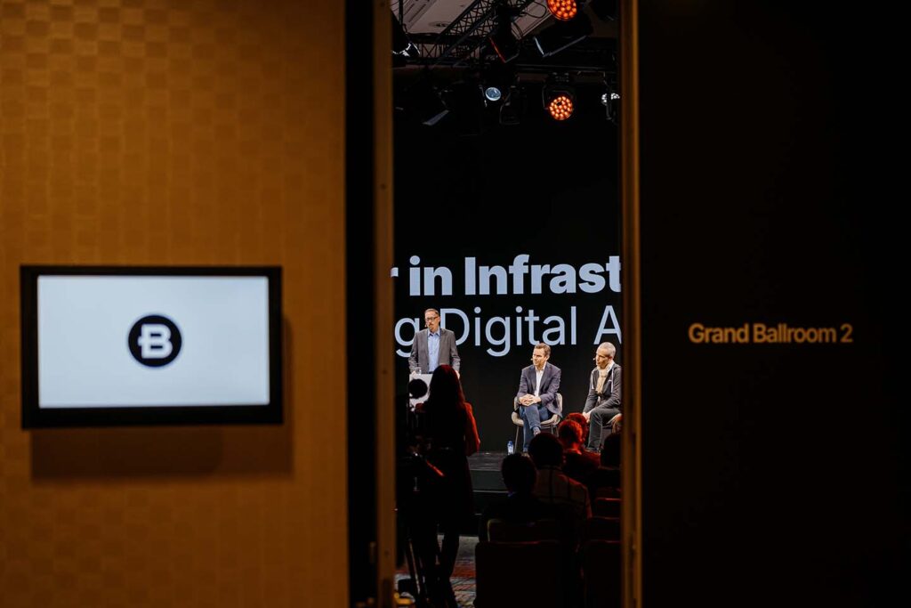 A panel of three speakers sits on stage in front of an audience in Grand Ballroom 2, as seen through an open doorway.