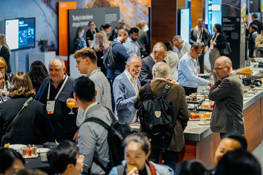 A large group of people at a conference networking and gathering around a buffet table with food and drinks in a modern indoor venue.