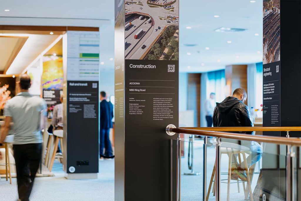 People interact in a modern indoor space with informational construction display panels and seating areas, viewed in natural daylight.