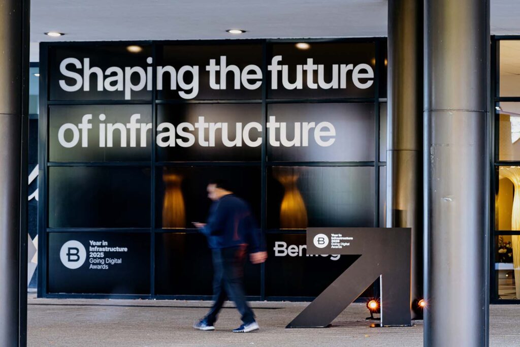 A person walks past a large glass wall displaying the text "Shaping the future of infrastructure" at an event venue.