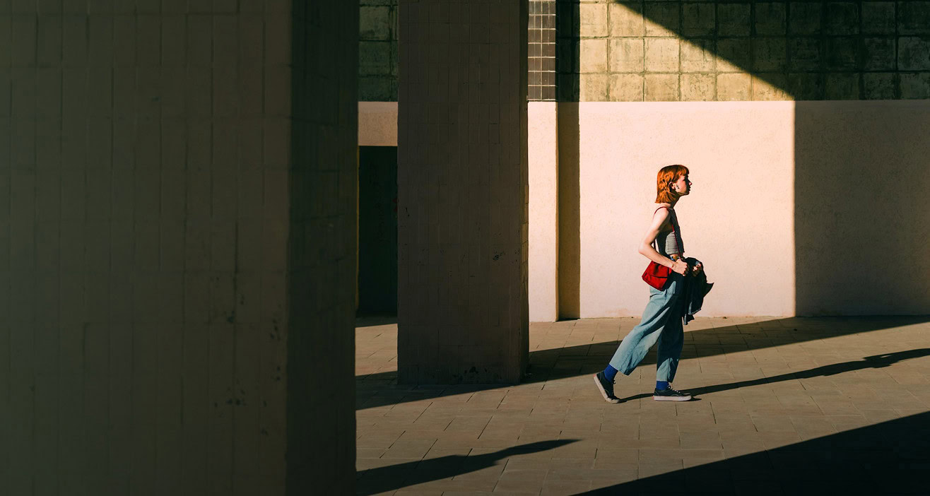 A person with red hair walks alone between large concrete columns in a sunlit, open urban space, casting a long shadow.