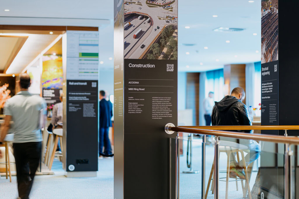 Indoor event space with informational panels on construction, people walking and sitting, and a railing in the foreground.