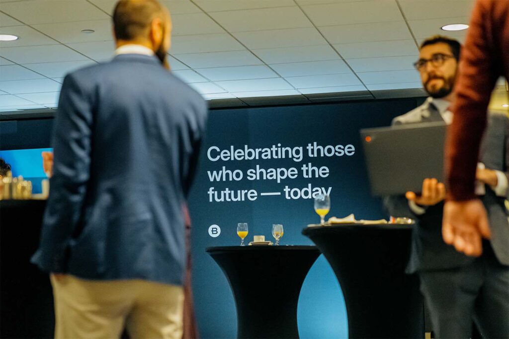 People in business attire stand near cocktail tables at an event. A wall displays the text, "Celebrating those who shape the future — today.